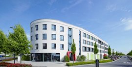 Ein modernes, kurvenförmiges Gebäude mit großen Fenstern und bunten Akzenten, umgeben von grünen Bäumen und einem blauen Himmel.