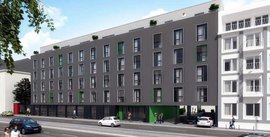 Ein modernes, mehrstöckiges Gebäude mit grauer Fassade und grünen Akzenten, umgeben von einer urbanen Umgebung.