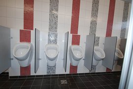 Die Innenansicht einer Herrentoilette zeigt mehrere Urinale, umgeben von grauen Trennwänden und bunten Fliesenmustern.