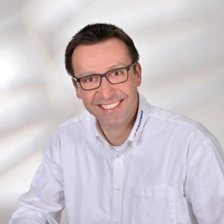 Professionelles Porträt eines lächelnden Mannes in Hemd Ein lächelnder Mann mit Brille trägt ein weißes Hemd und posiert vor einem hellen, verschwommenen Hintergrund.