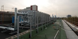 Auf dem Dach sind massive Kühleinheiten und Metallrohre zu sehen, während die Skyline im Hintergrund leicht verschwommen ist.