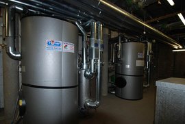In einem industriellen Raum stehen zwei große silberne Heizkessel, umgeben von zahlreichen Rohren und Ventilen.