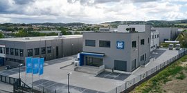 Ein modernes Firmengebäude mit grauer Fassade und blauen Akzenten, umgeben von einer gepflegten Außenanlage.