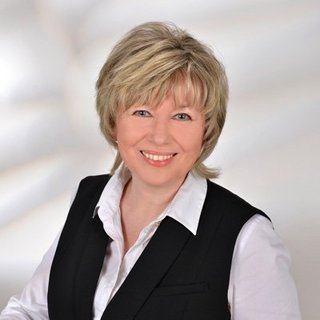 Professionelles Porträt einer lächelnden Frau in Anzug Die Frau mit blonden Haaren strahlt Freundlichkeit aus, während sie in einem eleganten Outfit vor einem sanften, neutralen Hintergrund steht.