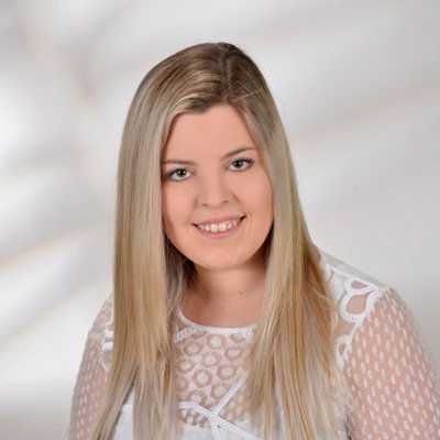 Eine junge Frau mit langen, blonden Haaren lächelt freundlich in die Kamera, während sie ein weißes, durchscheinendes Oberteil trägt.