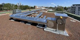 Die Installation von glänzenden Rohren auf einem Dach, umgeben von Kies, bietet einen Kontrast zur urbanen Umgebung und dem blauen Himmel.