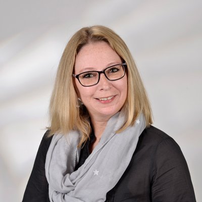 Eine Frau mit blonden Haaren und Brille lächelt freundlich in die Kamera, während sie einen grauen Schal trägt.