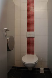 Die Innenansicht einer Toilette, die durch eine graue Tür zugänglich ist, mit einem auffälligen roten Fliesenstreifen in der Mitte.