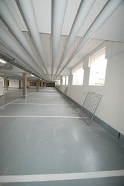 Ein weitläufiger Parkhausbereich mit grauem Boden und weißen Parkplatzmarkierungen, beleuchtet durch große Fenster.