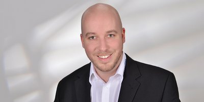 Der Mann mit einem freundlichen Lächeln hat einen gepflegten Bart und posiert in einem eleganten Outfit vor einem neutralen Hintergrund.