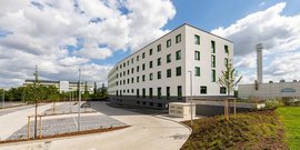 Ein modernes, mehrstöckiges Gebäude mit grünen Fenstern und einem gepflegten Außenbereich unter einem bewölkten Himmel.