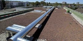 Eine Dachlandschaft mit silbernen Rohren, die sich über eine Kiesfläche erstrecken, umgeben von städtischen Gebäuden im Hintergrund.