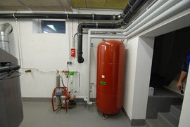 Ein roter Drucktank steht in einem modernen Heizungsraum, umgeben von Rohren und einer Pumpe auf dem Boden.