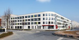 Ein modernes Wohngebäude mit geschwungener Fassade und großen Fenstern, umgeben von einer ruhigen Straße und Bäumen.