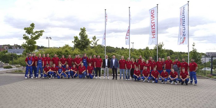 In der Mitte der Gruppe stehen zwei Männer in Anzügen, umgeben von Mitarbeitern, die stolz ihre Unternehmenskleidung tragen.