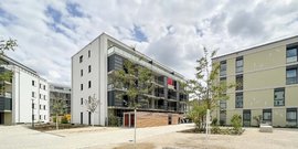 Moderne Wohngebäude umgeben von Bäumen und einem gepflasterten Platz, unter einem bewölkten Himmel.