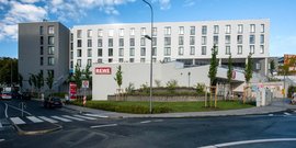 Ein helles, modernes Gebäude mit einem REWE-Supermarkt, das von einer grünen Landschaft und einer ruhigen Straße umgeben ist.