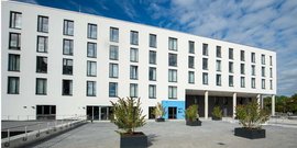 Die Architektur zeigt ein elegantes, helles Gebäude mit klaren Linien und einem blauen Eingang, unter einem bewölkten Himmel.