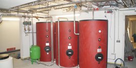 Der Raum zeigt drei rote Heizkessel, die an ein komplexes Rohrsystem angeschlossen sind, mit einem grünen Druckbehälter in der Ecke.