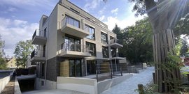 Ein modernes Wohngebäude mit großen Fenstern und Balkonen, umgeben von Bäumen und einer gepflasterten Einfahrt.