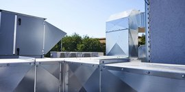 Eine Reihe von silbernen Lüftungskanälen auf einem Dach, umgeben von grünem Laub im Hintergrund unter klarem Himmel.