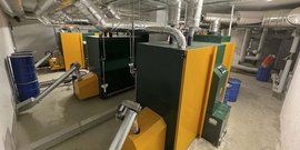 In einem Heizungsraum stehen mehrere große Heizkessel in gelb und grün, umgeben von Rohren und blauen Fässern.