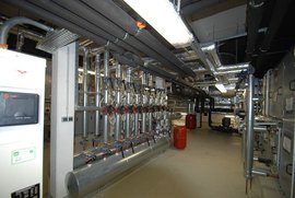 Die Umgebung zeigt eine Vielzahl von Metallrohren und Ventilen, die in einem industriellen Setting zur Steuerung von Flüssigkeiten dienen.