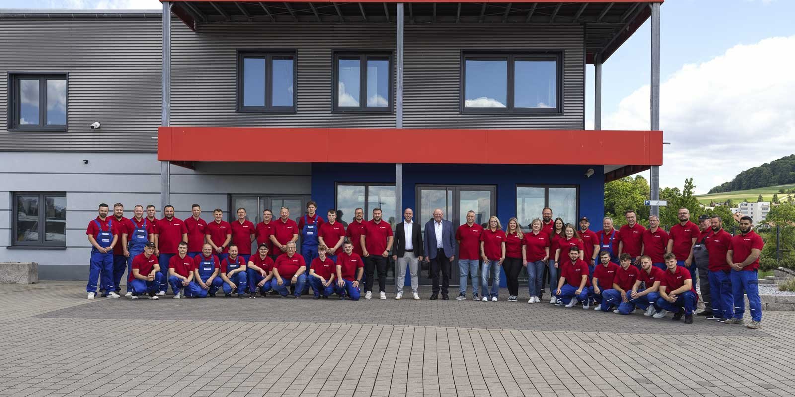 Mitarbeiter in einheitlicher Kleidung stehen in zwei Reihen vor einem Gebäude mit großen Fenstern und einem roten Vordach.