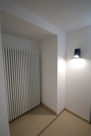 In einer Ecke eines minimalistischen Raumes steht ein schlanker Heizkörper, beleuchtet von einer eleganten Wandlampe.