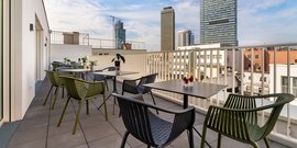 Eine moderne Dachterrasse mit eleganten Stühlen und Tischen, umgeben von urbanen Gebäuden und einem klaren Himmel.