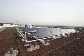 Auf dem Dach sind mehrere Solarpanels installiert, umgeben von Kies und kleinen Pflanzen, mit einem grauen Himmel im Hintergrund.