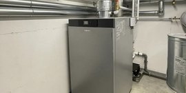 Ein moderner Heizkessel steht in einem Kellerraum, umgeben von silbernen Rohren und einer Metalltrommel.