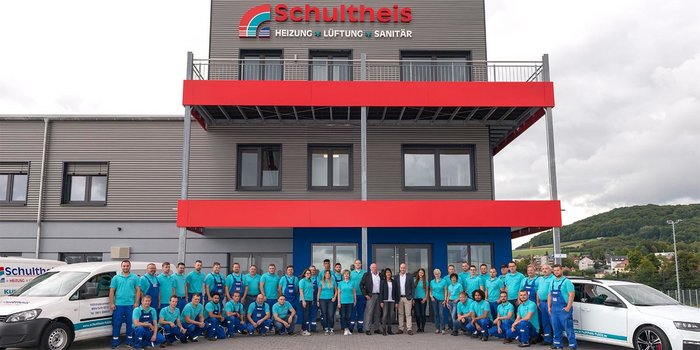Das Team von Schultheis posiert lächelnd vor ihrem neuen Bürogebäude, umgeben von Fahrzeugen und einer grünen Landschaft.