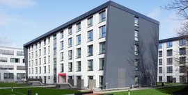 Ein modernes, mehrstöckiges Gebäude mit einer schrägen Fassade und großen Fenstern, umgeben von einer gepflegten Grünfläche.