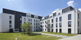 Die Architektur der Wohngebäude kombiniert helle und dunkle Elemente, während der Rasen und die Wege einladend wirken.