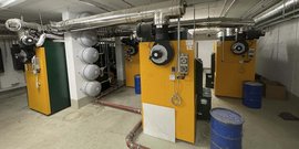 Ein industrieller Raum mit mehreren Heizkesseln in Gelb und Grün, umgeben von Rohren und blauen Fässern auf dem Boden.