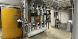 Ein großer gelber Wassertank steht in einem technischen Raum, umgeben von Rohren und Ventilen, die für die Heizungsanlage zuständig sind.