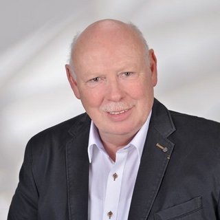 Professionelles Porträt eines lächelnden Mannes in Anzug Ein älterer Mann mit Glatze und Schnurrbart lächelt freundlich in die Kamera, gekleidet in einem dunklen Blazer und weißem Hemd.