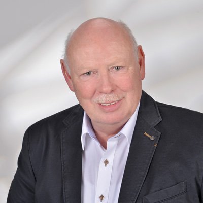 Ein älterer Mann mit Glatze und Schnurrbart lächelt freundlich in die Kamera, gekleidet in einem dunklen Blazer und weißem Hemd.