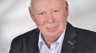 Ein älterer Mann mit Glatze und Schnurrbart lächelt freundlich in die Kamera, gekleidet in einem dunklen Blazer und weißem Hemd.