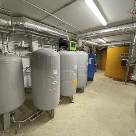 In einem technischen Raum stehen mehrere graue Druckbehälter, umgeben von Rohren und einer gelben Tankanlage.